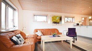 A living room with a brown leather sofa, a wooden coffee table, and a purple chair. There are two windows with curtains, a painting on the wall, and a kitchen area with a microwave.