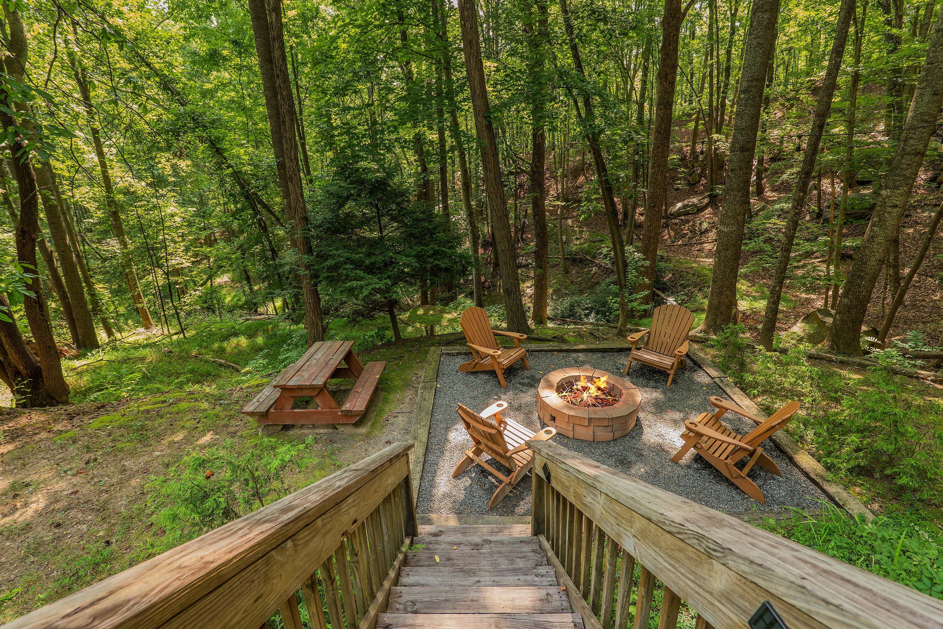 A forested area with a wooden deck leading to a fire pit surrounded by Adirondack chairs.