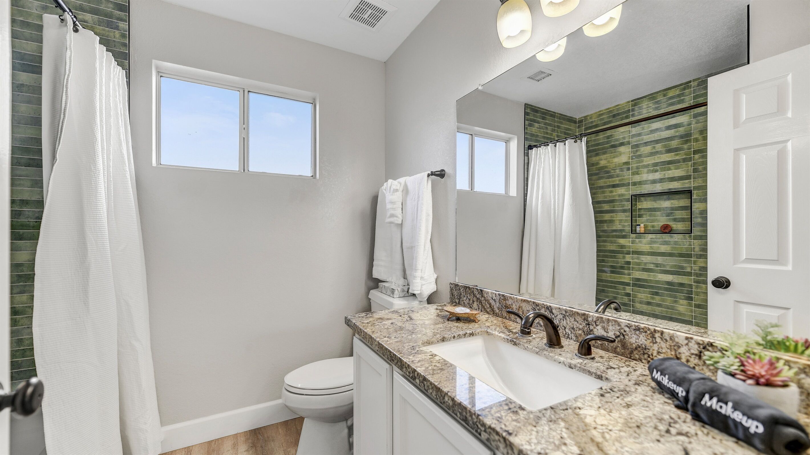 Une salle de bain moderne avec des toilettes blanches, un lavabo doté d’un plan de travail en granit, un grand miroir et une douche avec un rideau.