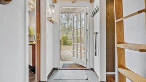Um corredor com porta de madeira que dá para um jardim, um tapete pequeno e um candeeiro fixado na parede.
