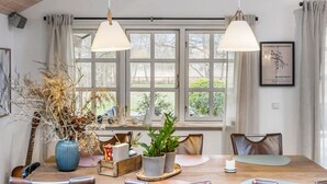 Uma área de refeições com mesa de madeira, cadeiras, luminárias pendentes e uma janela com vista para árvores.