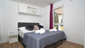 A neatly made bed with pillows and towels, a bedside table with a lamp, a window with curtains, and a white shelf with a plant.