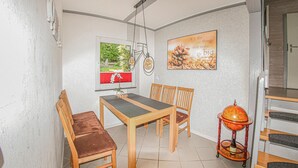 Un comedor con una mesa y sillas de madera, una ventana con persiana roja, una lámpara de pared y una cita inspiradora enmarcada.