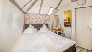 Un dormitorio con cabecera de piedra, cama con ropa de cama blanca, mesita de noche con lámpara y puerta con luz circular.
