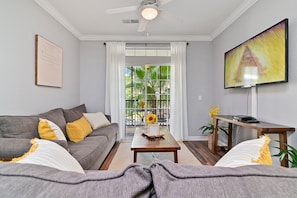 A living room with a grey sofa, a coffee table with a vase, a flat-screen TV, and a window with curtains.