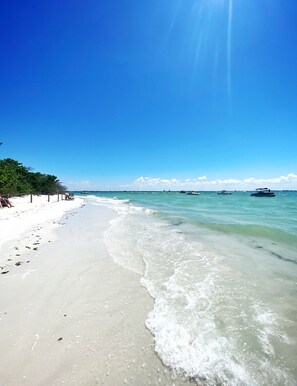 A beach with clear turquoise water, a sandy shore, and a few boats in the distance.