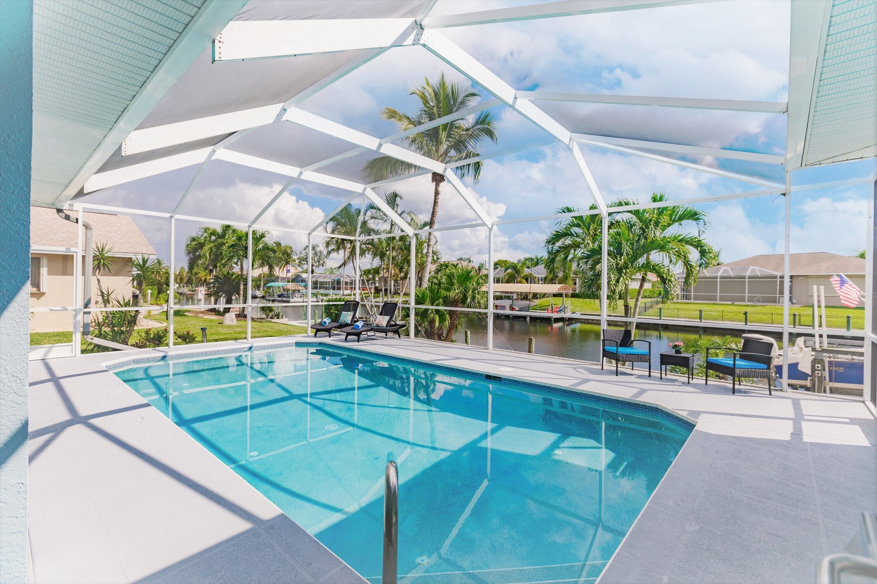 A swimming pool with a covered area, lounge chairs, and a view of palm trees and a body of water.