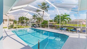 Una piscina con area coperta, poltrone prendisole e vista su palme e uno specchio d'acqua.