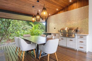 An outdoor kitchen with a grill, dining table, and chairs, surrounded by a wooden deck and greenery.