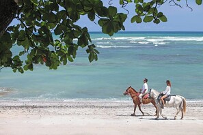 Two individuals riding horses on a beach with ocean in the background.