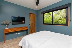 A bedroom with a bed, a TV mounted on the wall, a wooden bench, a door, and a window with a view of greenery.