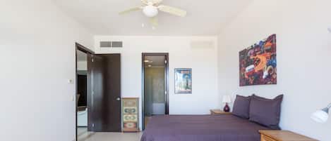 A bedroom with a bed, a ceiling fan, a wooden cabinet, a nightstand with a lamp, and a poster on the wall.