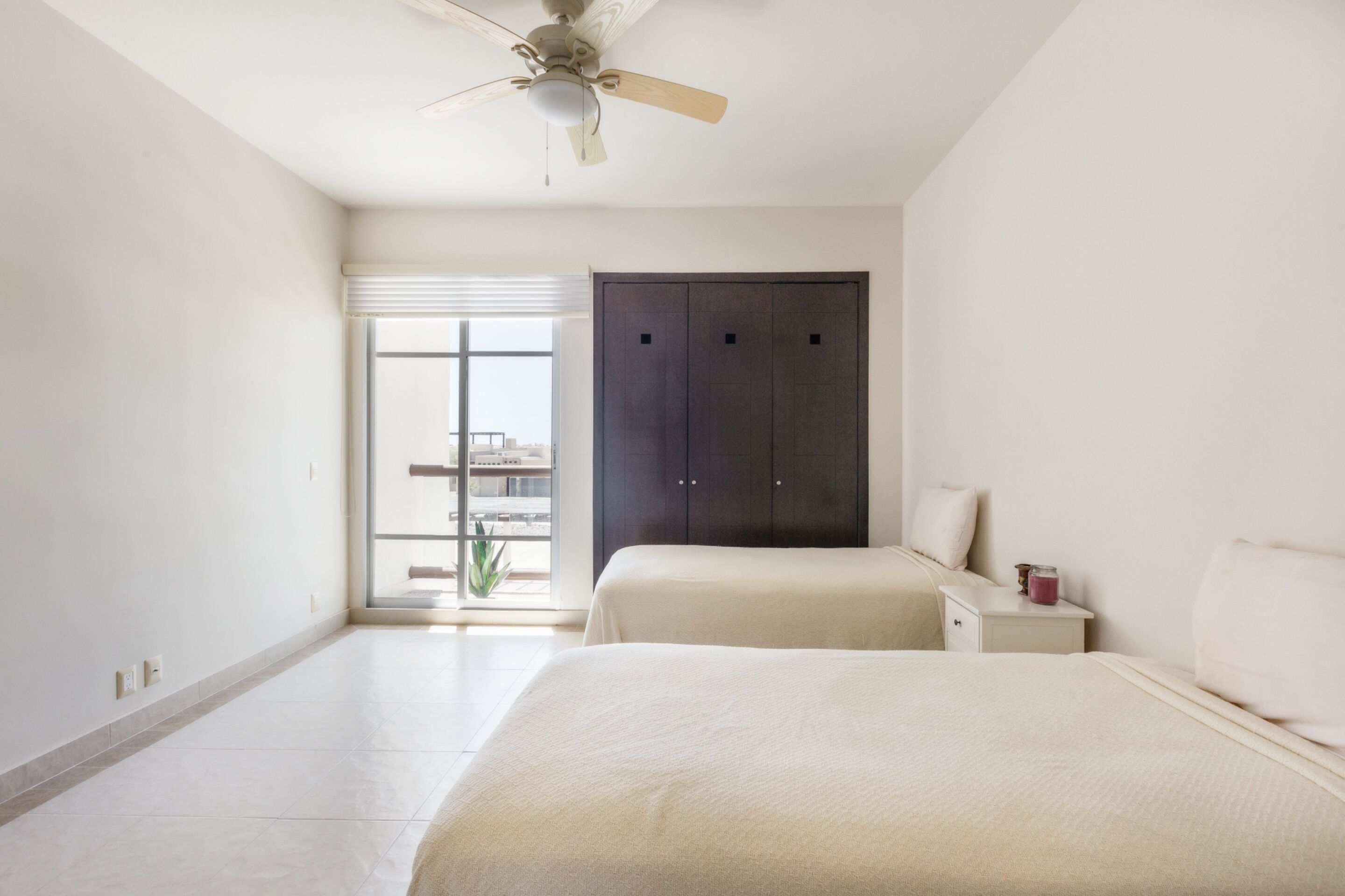 A bedroom with two beds, a ceiling fan, a dark wardrobe, a nightstand, and a window with a view of buildings.