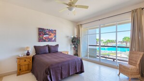 Un dormitorio con cama, mesita de noche, lámpara, ventilador de techo, ventana con vista a una piscina y una silla.