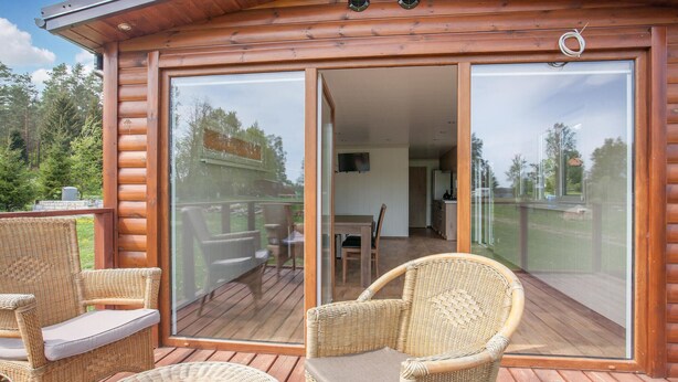 Une terrasse en bois, meublée de meubles en osier, et une porte-fenêtre coulissante donnant sur un espace intérieur comprenant un coin repas et une cuisine.