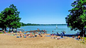 Une plage avec une jetée en bois, des gens qui prennent un bain de soleil et des bateaux sur l’eau.
