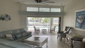 Ein geräumiges Wohnzimmer mit Deckenventilator, großem Fenster und Blick auf Pool und Palmen.