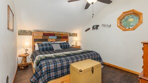 Un dormitorio con cama, ventilador de techo, mesita de noche, lámpara, cabecera de madera, decoración en la pared y un baúl para guardar objetos.