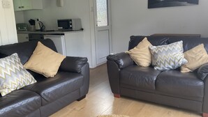 A living room with two leather sofas, a kitchen in the background, and a painting on the wall. - Holiday Cabin Sleeps 5 Caernarfon, Snowdonia (Caernarfon)