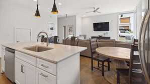 Cozinha moderna com ilha central, armários brancos e lava-louças. Há uma área de jantar com mesa e cadeiras de madeira redondas. Uma TV de tela plana está montada na parede, e um ventilador de teto é visível.