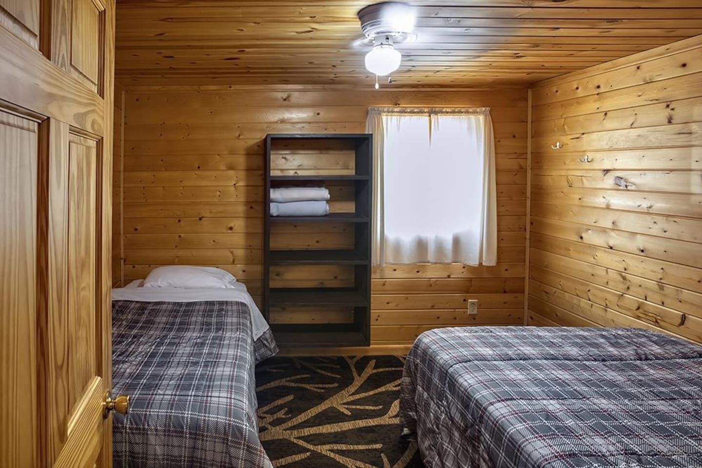 A small, cozy room with wooden walls, a bed with a plaid bedspread, a shelf with folded towels, and a window with a white curtain.