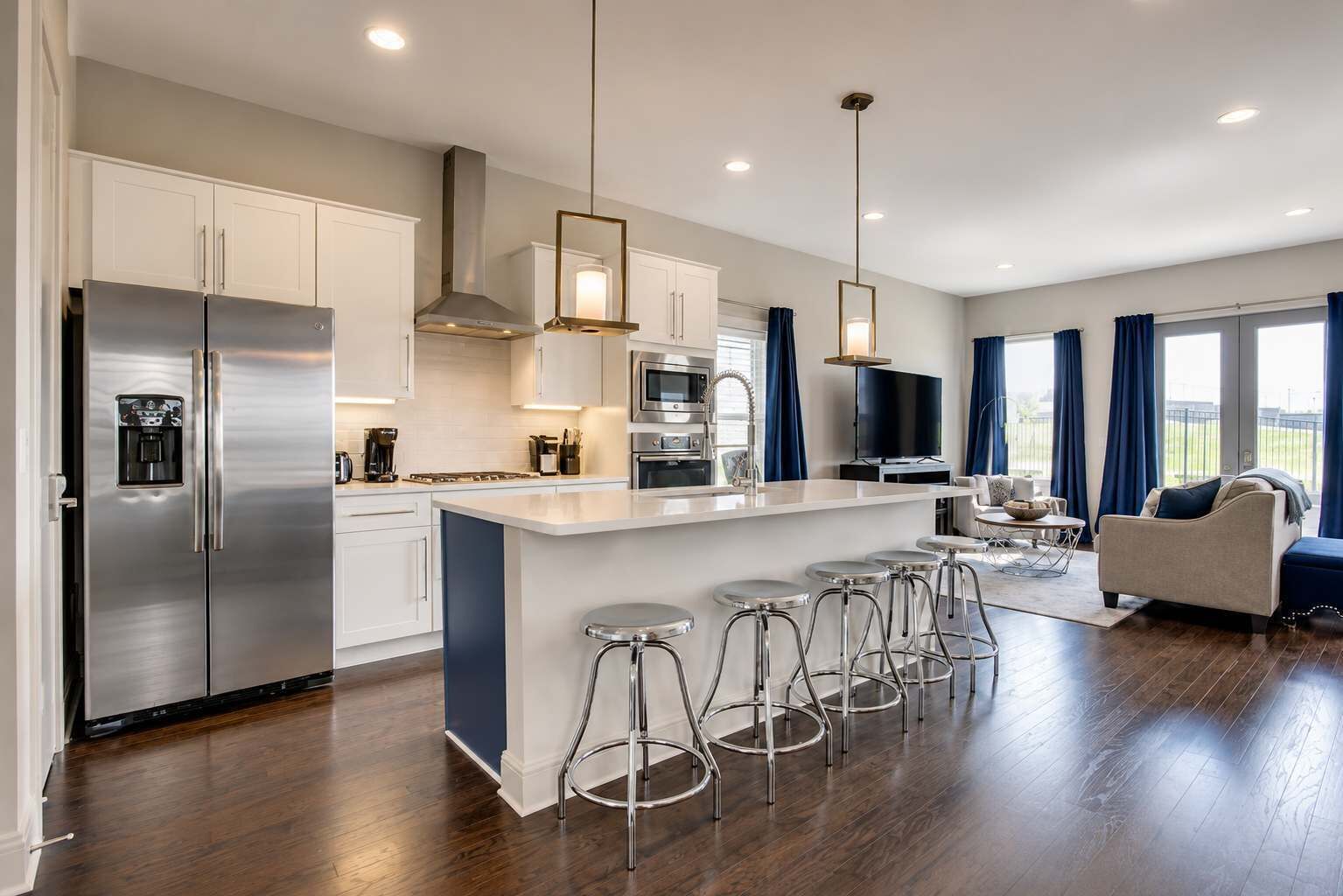 A modern kitchen with stainless steel appliances, a central island with bar stools, and a dining area with a sofa and TV.