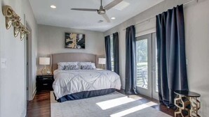 A bedroom with a large bed, a ceiling fan, a nightstand, a lamp, and a view of the outdoors through a window with curtains. - Stunning Nashville Retreat - Near Broadway (Nashville)