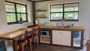 Une cuisine compacte avec des plans de travail en bois, un évier et un four à micro-ondes. Deux fenêtres laissent entrer la lumière naturelle, et une petite étagère est placée au-dessus de l’évier.