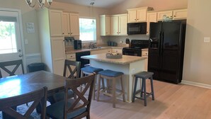 A modern kitchen with white cabinets, a black refrigerator, a microwave, and a coffee maker. There is a dining table with chairs and a small island with stools. - Sea You Soon- 1 mile to Camp Lejeune! Monthly rental- fully furnished!  (Hubert)