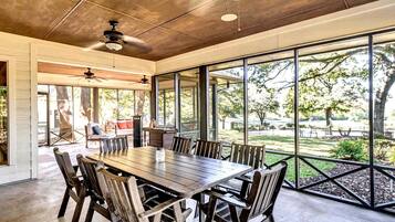 A spacious dining area with a large wooden table and chairs, large windows offering a view of the outdoors, and ceiling fans.
