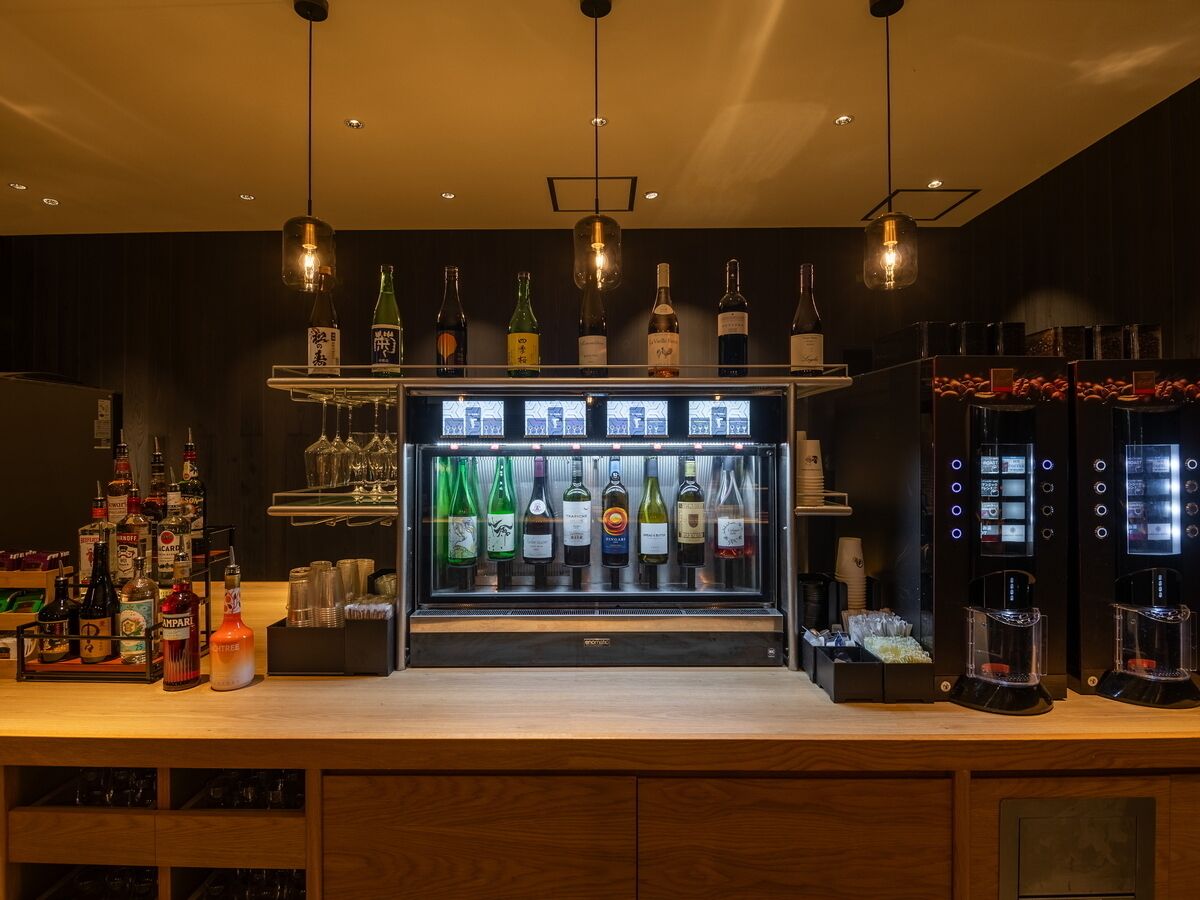 Un espace bar comprenant un présentoir réfrigéré où sont exposées différentes bouteilles, une machine à café et un assortiment de verres.