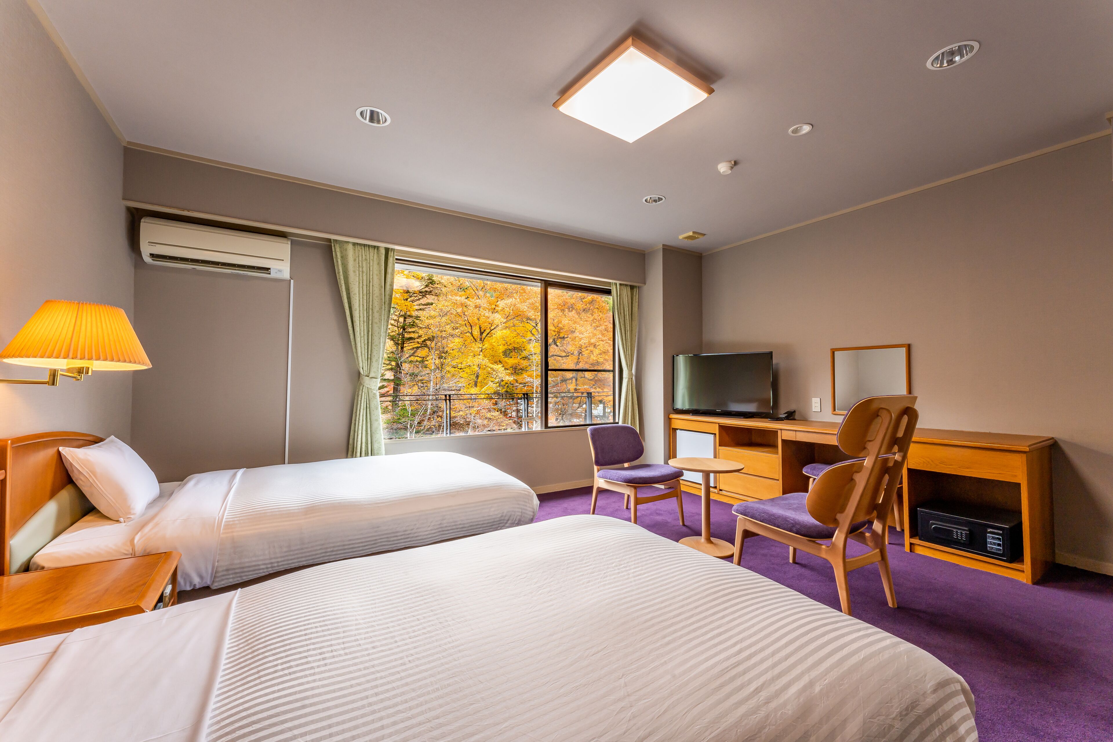 A hotel room with two beds, a desk with a chair, a television, and a window with autumn foliage view.