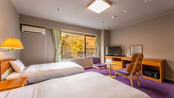A hotel room with two beds, a desk with a chair, a television, and a window with autumn foliage view.