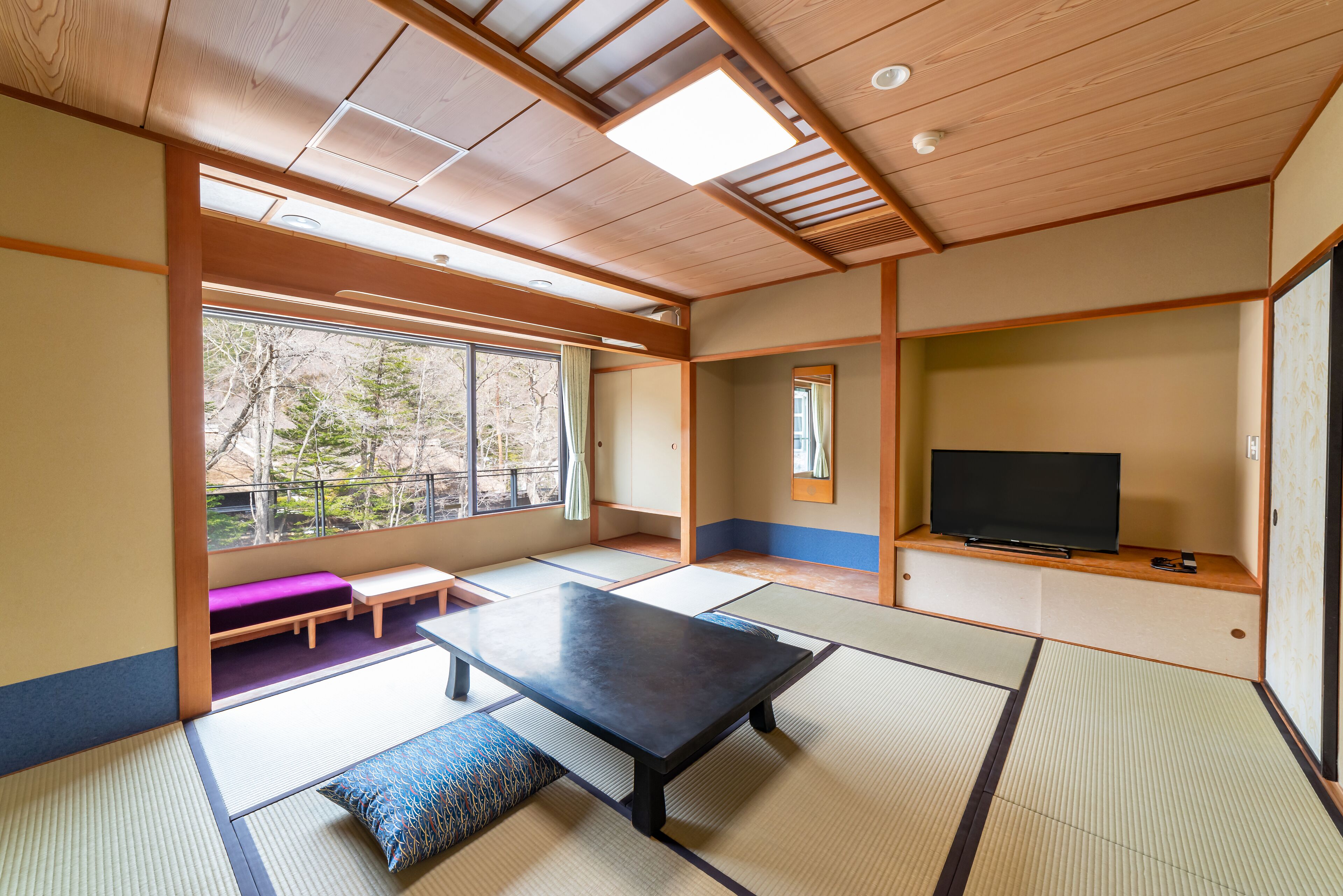 A traditional Japanese room with tatami flooring, a tatami mat table, a television, and a view of trees through large windows.