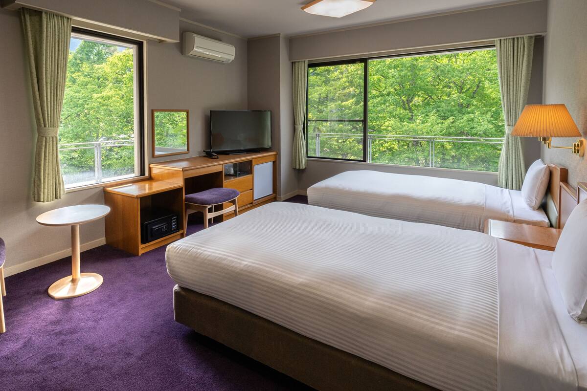 Habitación de hotel con dos camas, un escritorio con televisor, una mesita pequeña y ventanas amplias con vistas a árboles.