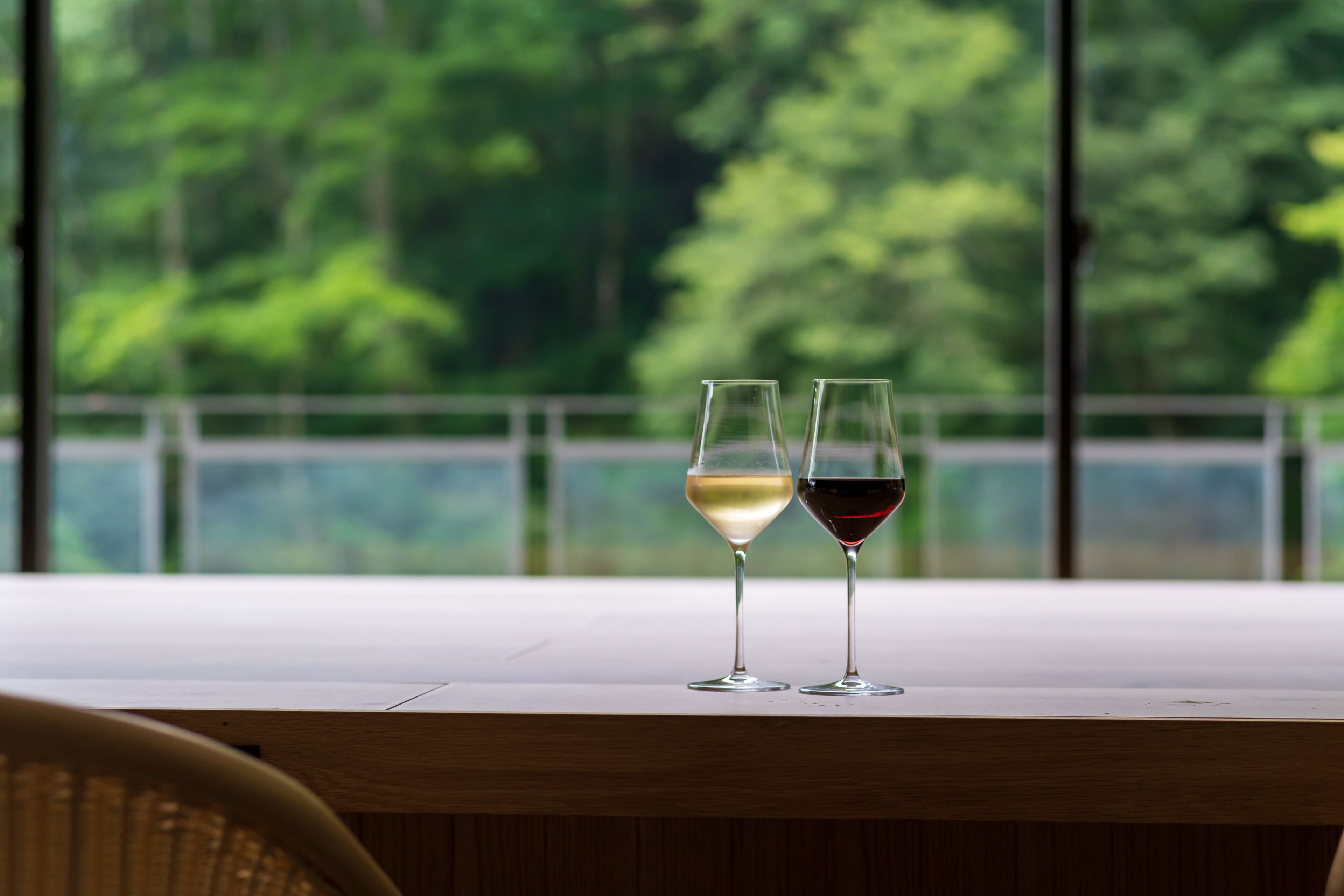 Two glasses of wine on a table with a view of trees through large windows.
