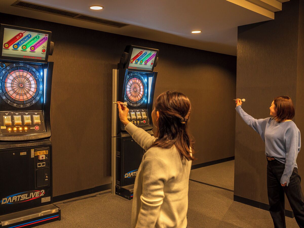 Two people playing darts at a Dartsline 2 machine in a room with dark walls and carpet.