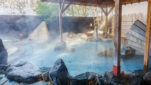 Une zone de sources chaudes surmontée d’une structure en bois, de la vapeur s’élevant de l’eau et des rochers entourant la piscine.