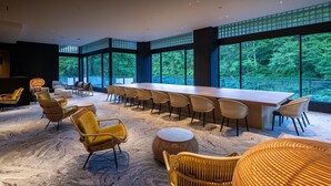 Une pièce spacieuse avec une longue table en bois et de nombreuses chaises, de grandes fenêtres donnant sur un paysage verdoyant, et un plafond haut doté d’un éclairage encastré.