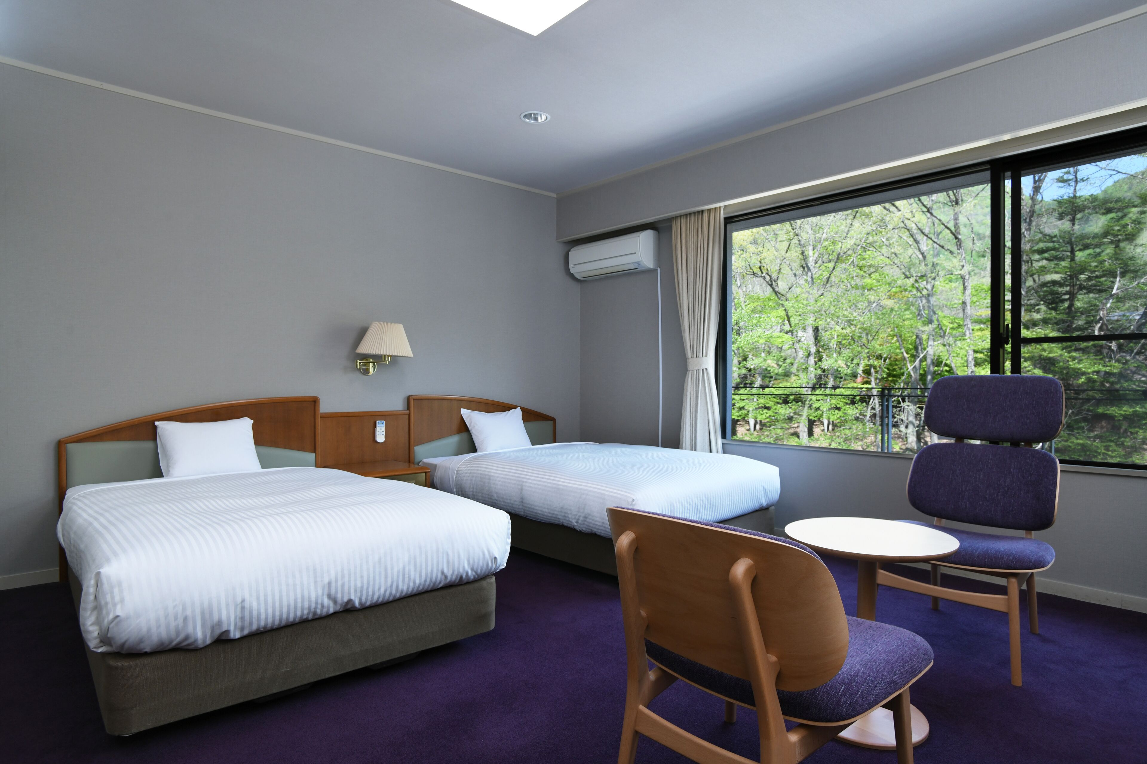 Een hotelkamer met twee bedden, een stoel, een klein tafeltje en een raam met uitzicht op bomen.