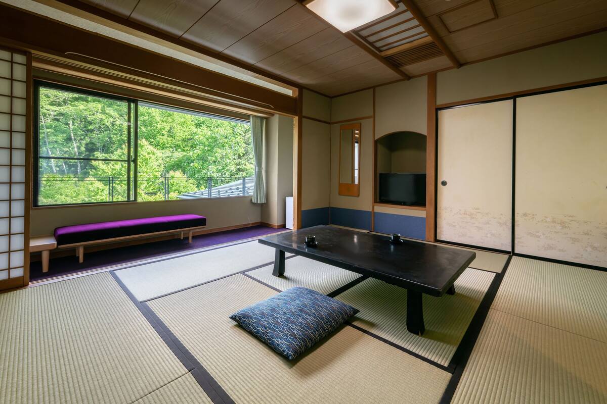 Una habitación con suelo de tatami, una mesa de tatami, un banco morado y un ventanal con vistas a árboles.