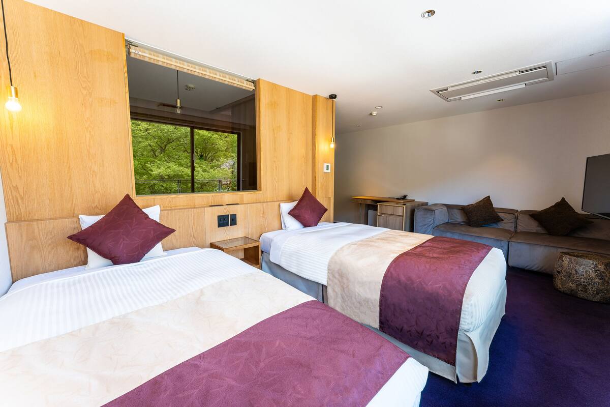 Habitación de hotel con dos camas, un sofá, un televisor y ventana con vista a árboles.