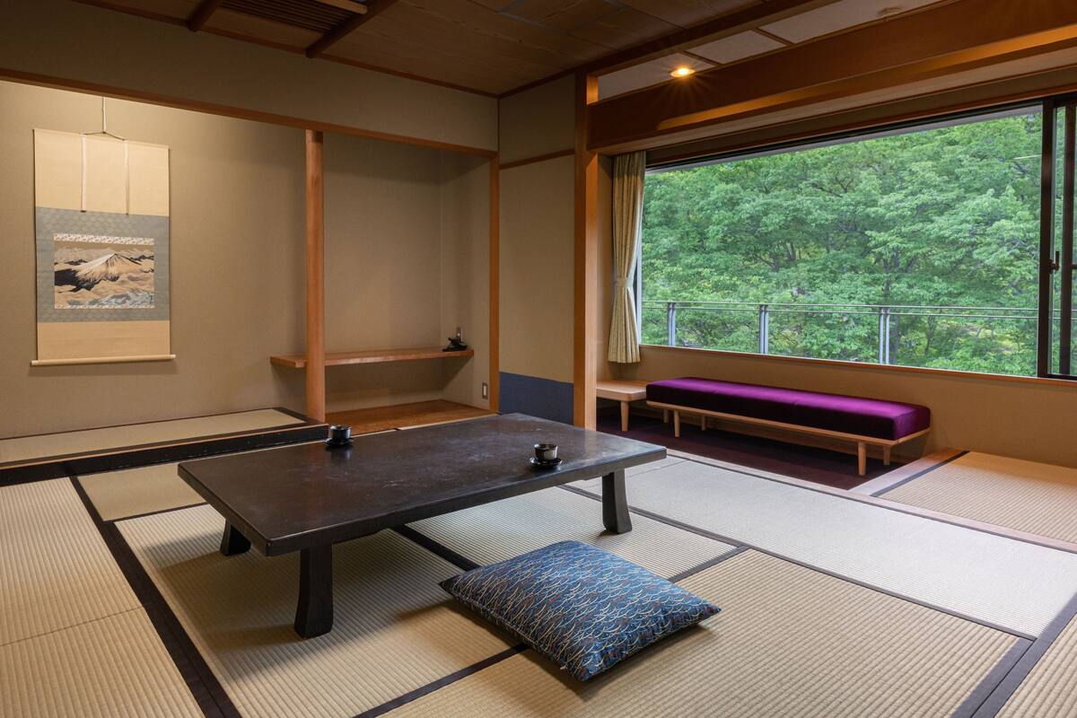Una habitación con suelo de esteras de tatami, una mesa de tatami, un cojín púrpura, un estante de madera y vistas a vegetación a través de una ventana.