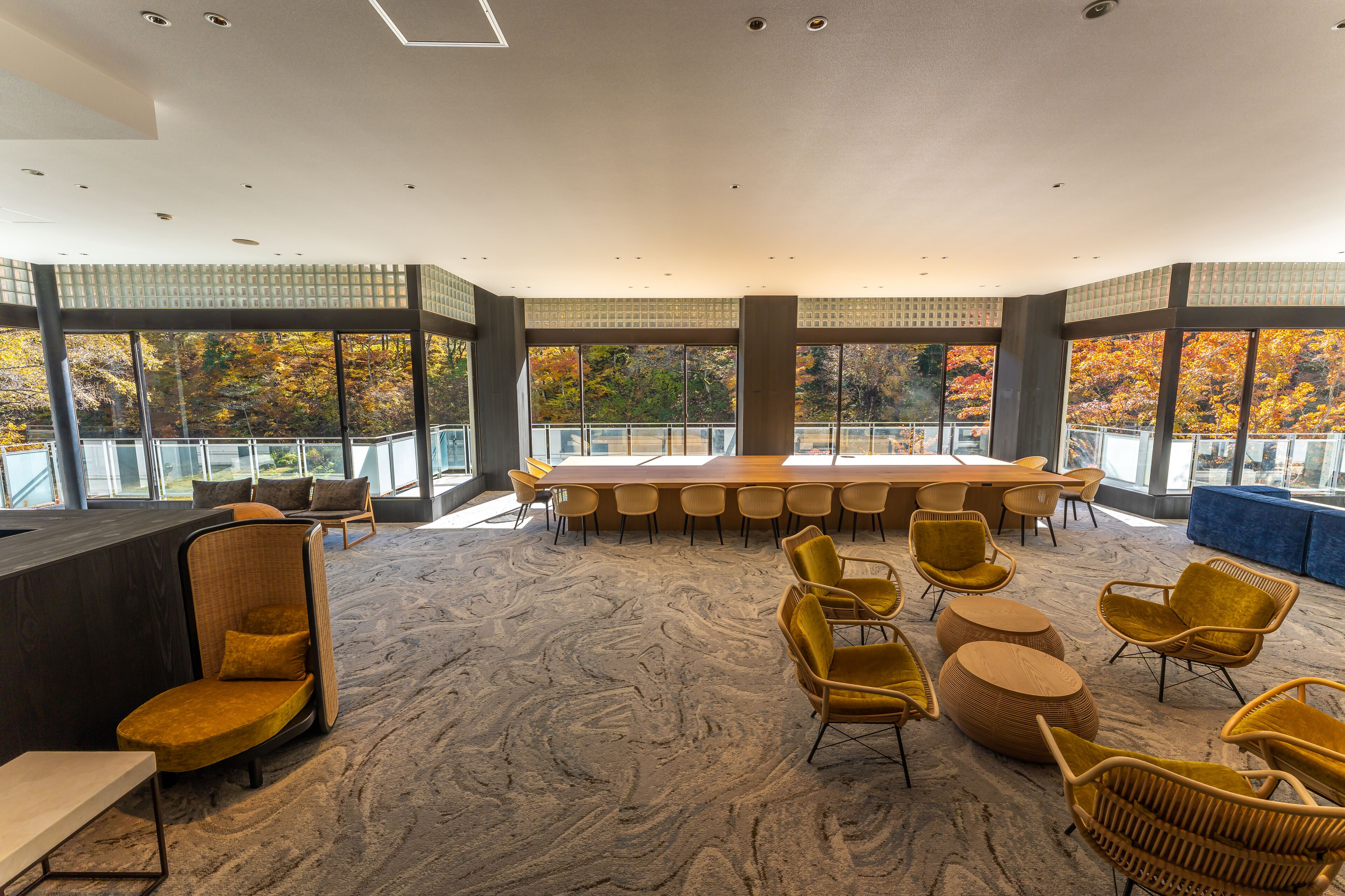 A spacious conference room with a large table and chairs, surrounded by large windows offering a view of autumn foliage.