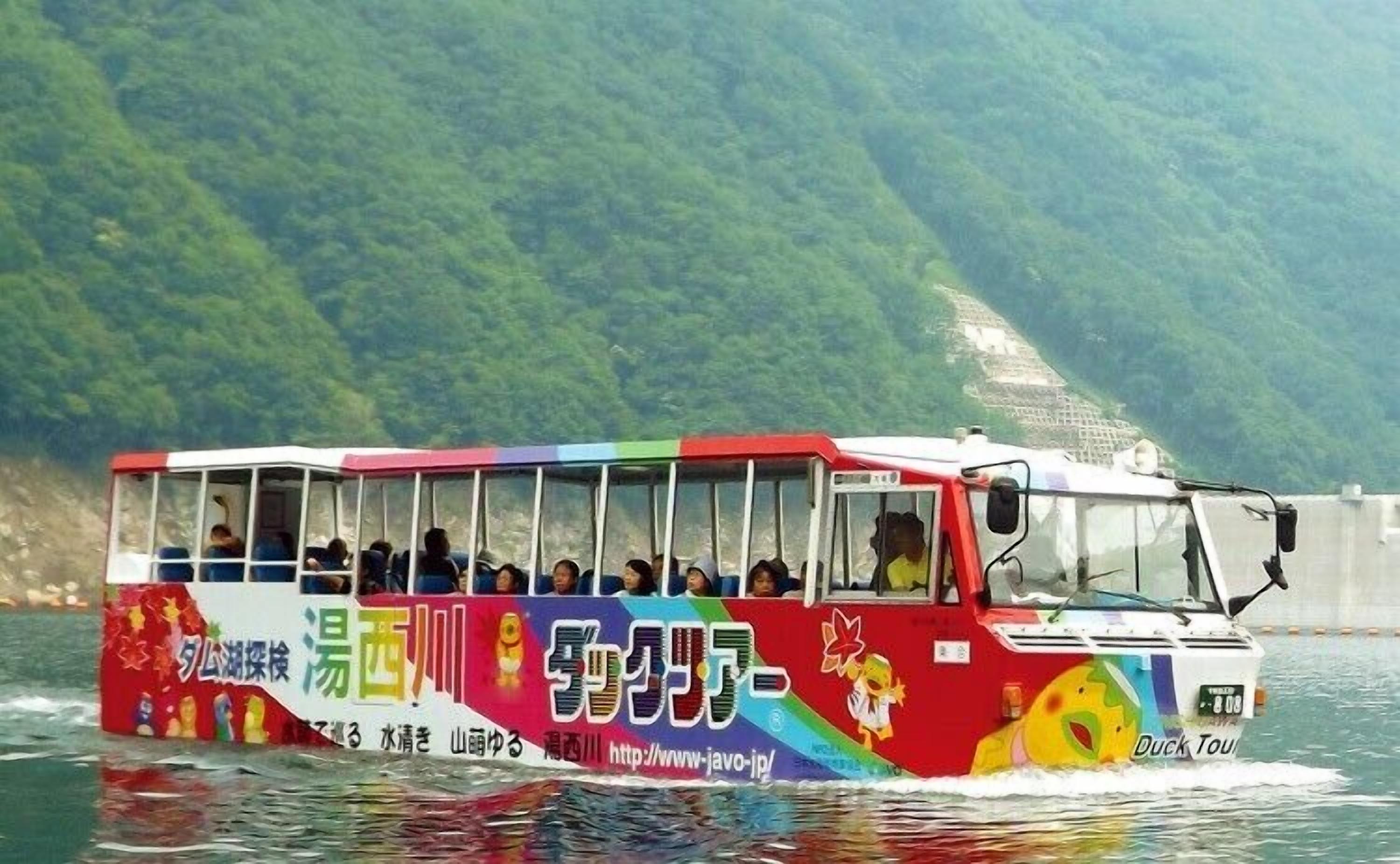 Un autobús colorido con pasajeros viajando sobre el agua, con montañas al fondo.