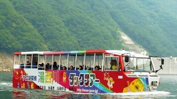 Un autobús colorido con pasajeros viajando sobre el agua, con montañas al fondo.