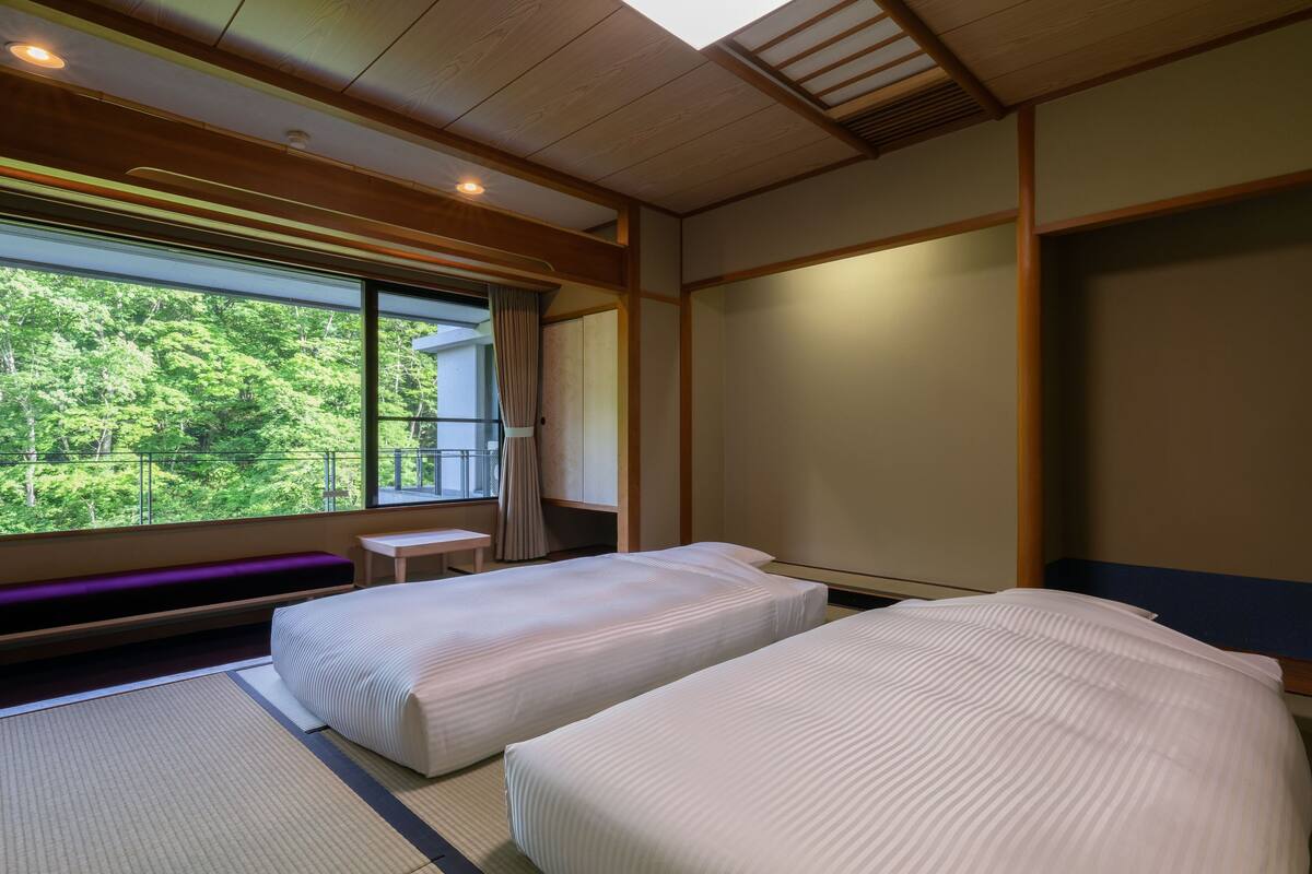 Habitación de hotel con dos camas, una mesita pequeña y vistas a la vegetación a través de amplias ventanas.