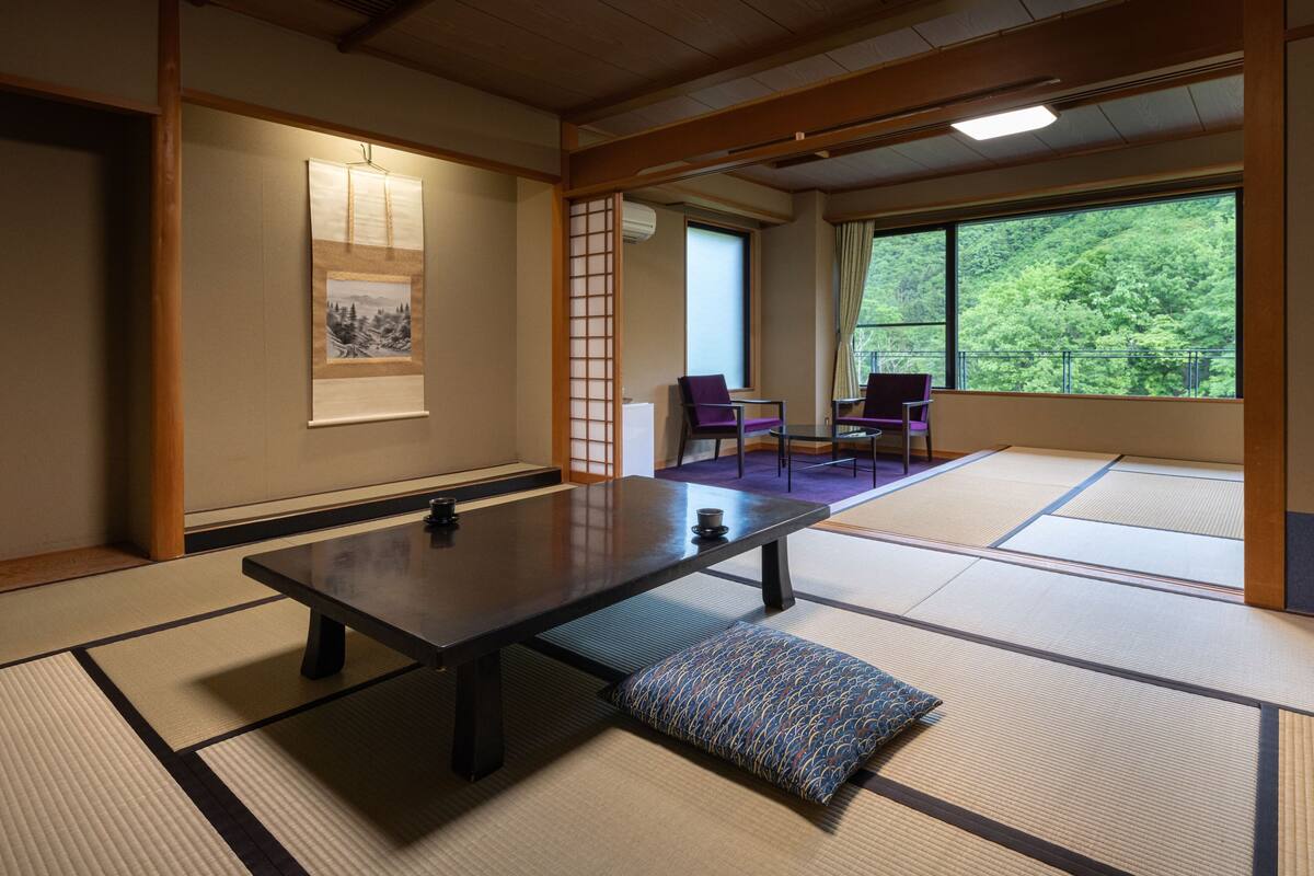 Una habitación japonesa tradicional con suelo de tatami, una mesa baja, dos sillas y vistas a un bosque a través de amplios ventanales.