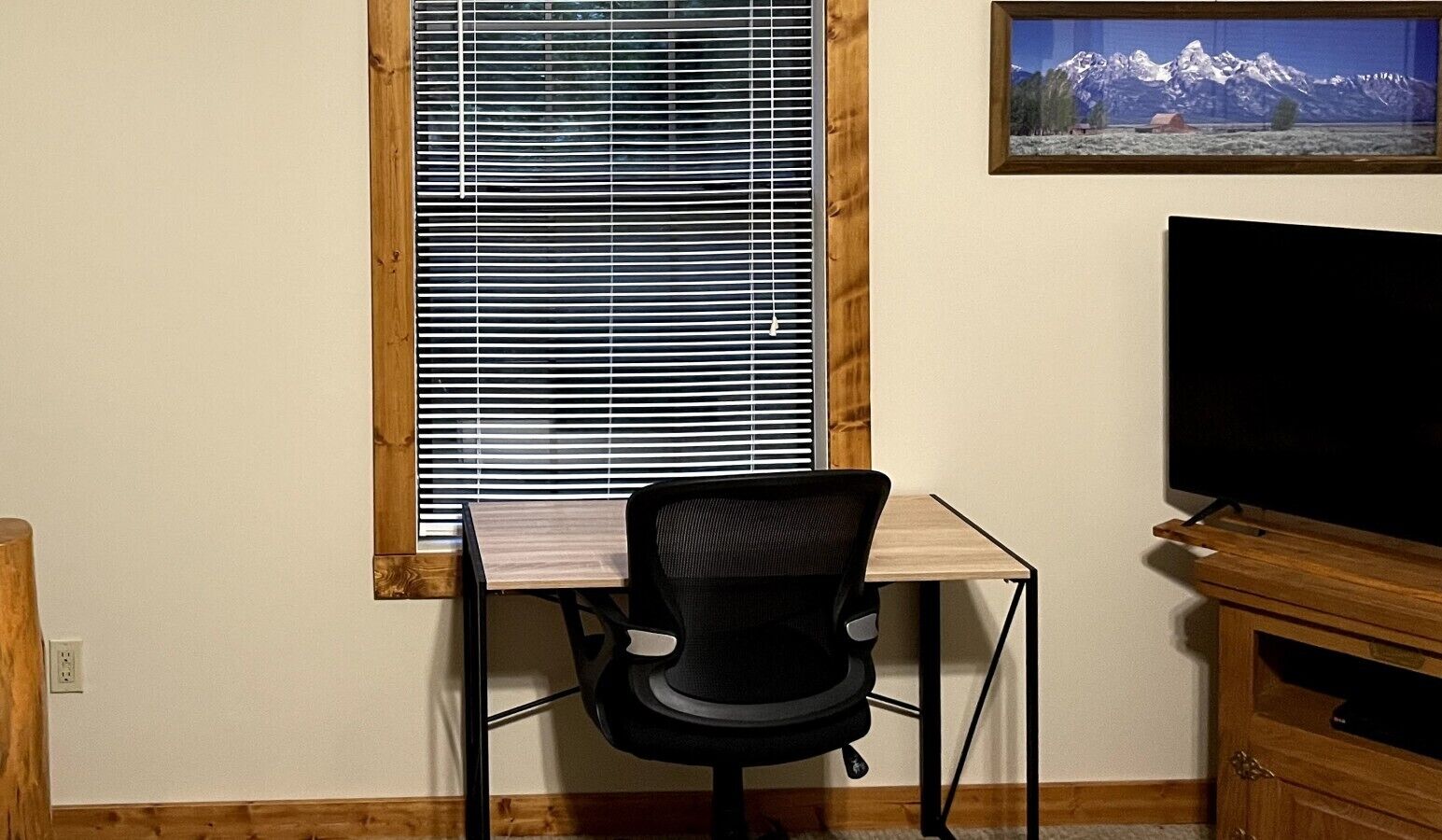 Une pièce comprenant un bureau, une chaise, un téléviseur et une fenêtre avec des stores.