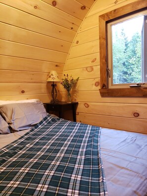 Ein gemütliches Schlafzimmer mit Holzbalkendecke, einem Fenster mit Blick auf Bäume, einem Nachttisch mit Lampe und Blumenvase sowie einem Bett mit kariertem Überwurf.
